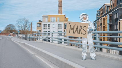 Man in Spacesuit is Standing at the Edge of a Road and Holding a Sign with Mars Written on it....