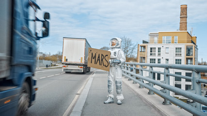 Man in Spacesuit is Standing at the Edge of a Road and Holding a Sign with Mars Written on it....