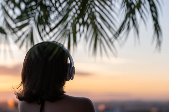 Back view silhouette of relaxed woman wearing headphones meditating listening to music on the beach at sunset in the branches of palm trees. - Powered by Adobe