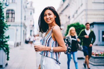 Half length portrait of cheerful asian woman dressed in trendy outfit standing with coffee to go on square in downtown, attractive female tourist satisfied with getting to showplace looking at camera