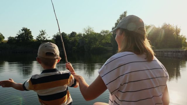 Happy mom teaches little son to fish while sitting on a wooden pier by the lake on a sunny summer day