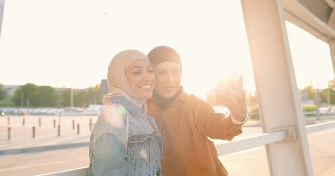 Two beautiful young Arabian women in headscarves smiling and taking selfie photos outdoors. Pretty cheerful muslims females posing to mobile phone and making photo. Girls with smartphone at street.