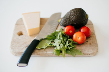 Avocado, cherry tomato, arugula, parmesan and knife on wooden board on white modern kitchen. Process of making home toast with avocado. Home cooking concept