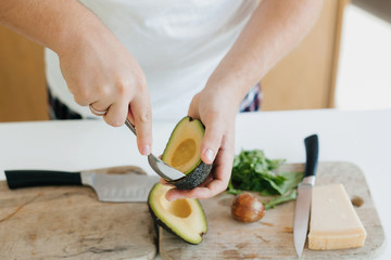 Person peeling perfectly ripe avocado with spoon for sandwich on modern white kitchen. Process of making healthy toasts with avocado, tomato, arugula, cheese. Home cooking concept