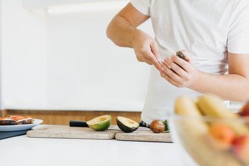 Person peeling perfectly ripe avocado with spoon for sandwich on modern white kitchen. Process of making healthy toasts with avocado, tomato, arugula, cheese. Home cooking concept