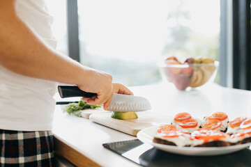 Person cutting avocado for sandwich on modern white kitchen. Process of making healthy toasts with avocado, tomato, arugula, cheese and whole grain bread. Home cooking concept