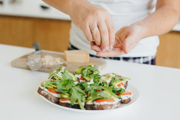 Hands putting parmesan on homemade toasts with avocado on modern white kitchen. Sandwich made of whole grain bread, avocado, tomato, arugula and cheese. Home cooking concept