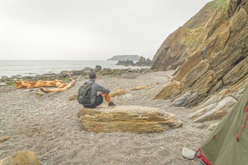 Obraz premium Male person with backpack sits on the roack and watching the sea with green tent on the left. Hidden gems and wil camping in ireland