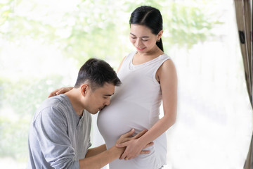 Asian husband is kiss his beautiful pregnant wife with love and they smiling together with happiness moment, concept of love, relation, expectation and  parenthood in  family lifestyle.