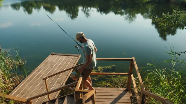 Dad and his little son with fishing rods in their hands go down the steps to a wooden pier to fish on the lake