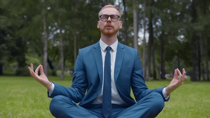 Relaxed businessman sitting on lawn doing meditation in city park