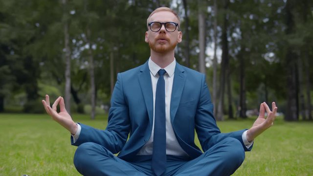 Relaxed businessman sitting on lawn doing meditation in city park