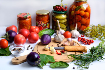 Cutting board with slices of garlic hot pepper, knife and eggplant, tomatoes, seasonings, herbs and jars of vegetables in the background on a light background.