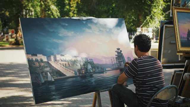 Man artist painting marine landscape with sailing ship and city on coast while sitting in park