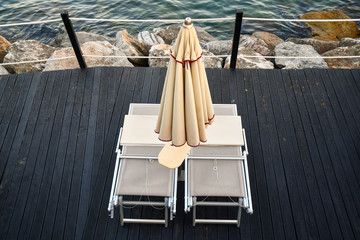 High-angle view of a pair of sunbeds under a closed beach umbrella on a wooden platform on the rocky sea shore, Liguria, Italy