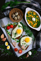 Two toasts with fried eggs on the board, tomatoes, appetizer of corn and peas, fork, garlic, quail eggs on a dark background