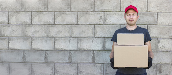 a young man delivery man stands against the background of a gray stone wall, holds two cardboard...