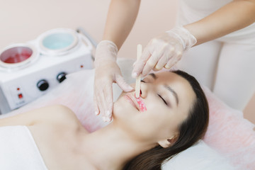 A master applies depilatory wax to the face of a beautiful young woman to remove unwanted hair....