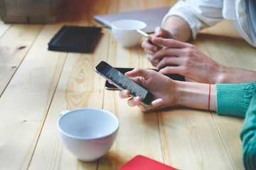 Cropped image of male's and female's hands installing mobile application and sharing information in social networks via modern digital cellulars with 4G connection sitting at wooden table with coffee