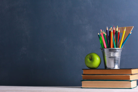 Fresh apple and colorful pencils pot on stacked books, school supplies on white desk with dark blue wall in background. Side view, copy space, close-up. Learning, education concept