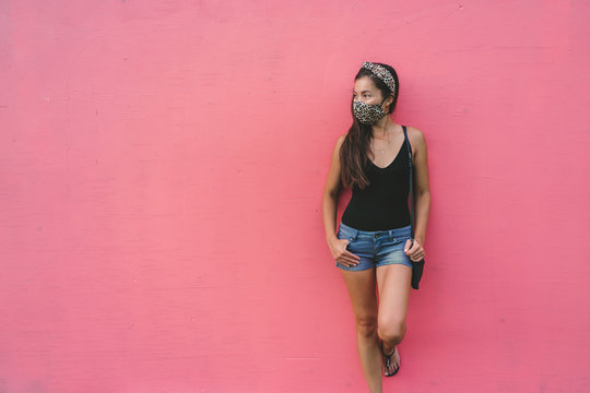 Asian woman leaning on pink wall wearing fabric face mask in fashion leopard pattern print. Casual young people city lifestyle during corona virus.