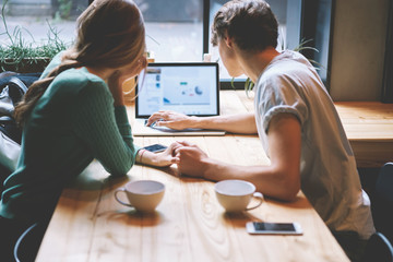 Obraz premium Back view of young couple in love holding each other hands while looking at macbook laptop monitor with copy space area for your advertising content.Hipsters resting in cozy cafe sitting at table