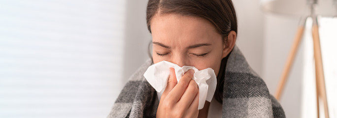 Sick woman feeling unwell staying home. Young girl with flu symptoms coughing in tissue covering nose when sneezing as COVID 19 prevention. Panoramic crop. - obrazy, fototapety, plakaty