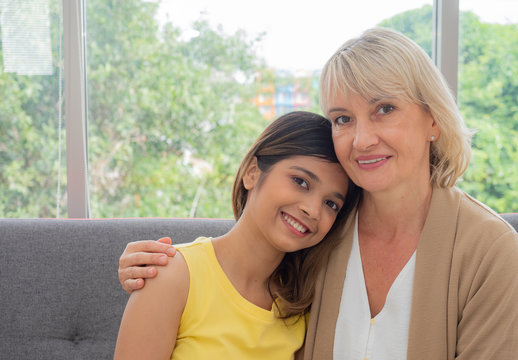 The adopted caucasian mother and Asian teen daughter happily hugged and smiled on the sofa. love and sharing time of stepmother, stepdaughter concept