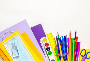 The concept of returning to school in a new format with a medical mask and disinfectant. Colorful school supplies on a white background. Space for text. The view from the top