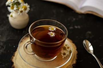English tea with chamomile stands on a wooden frame and a teaspoon and an open book for reading lies on a black background. Nearby is a white vase with summer chamomile flowers