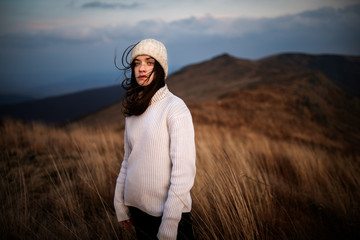 Young beautiful woman in a white sweater stands on top of a mountain and enjoys the scenery. Young woman standing on cliff's edge and looking to a sky - 371493413