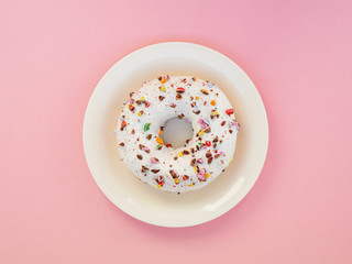 Homemade circle donut with white icing and choclate rainbow sprinkles on a trendy pink background and white plate situated in the center of image. Tasty doughnutdonut, top view. Sweet food leftlovers