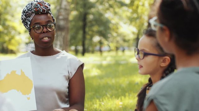 African American female teacher holding paper map and answering questions of school kids during outdoor lesson on summer day in the park
