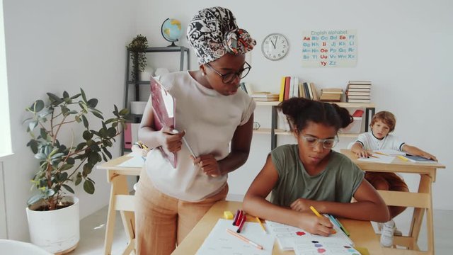 Helpful African American female teacher walking through classroom and talking to multiethnic kids while they drawing with felt tip pens in their workbooks at desks during primary school lesson