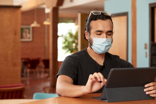 Young Hispanic man wearing black t shirt and clear frame glasses with face mask, sitting at bar in kitchen of downtown loft with iPad 
