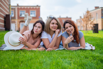 Three beautiful Caucasian young women lie on the lawn in the countryside. The blonde brunette and the redhead are resting in the park on the green grass. The friends had an outdoor picnic.