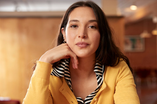 Portrait of young ethnic woman wearing yellow sweater with black and white striped blouse sitting at bar in kitchen of downtown loft 