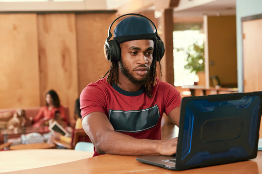Portrait of young ethnic man wearing red t shirt and knit hat wearing headphones, sitting at bar in kitchen of downtown loft with laptop computer 