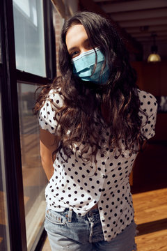 portrait of young ethnic woman with face mask wearing polka dot blouse and cut off jean shorts standing by bank of windows in downtown loft 