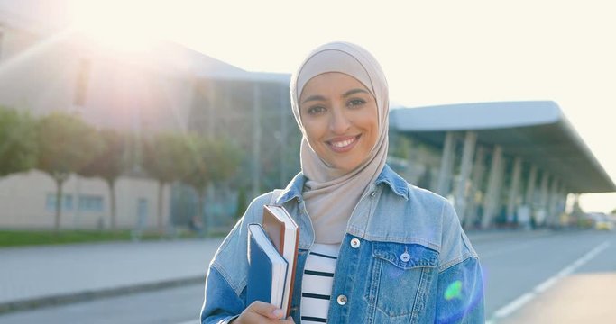 Portrait of young Arabic beautiful woman in headscarf smiling and holding textbooks and books. Pretty Arabian female student in hijab at street. Islamic happy girl. Education for Arabic girls.