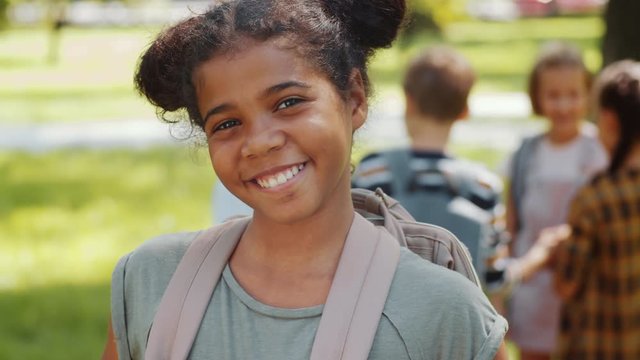 Chest up portrait shot of joyous African American school girl with backpack standing in park posing for camera and happily smiling while group of classmates talking in the background