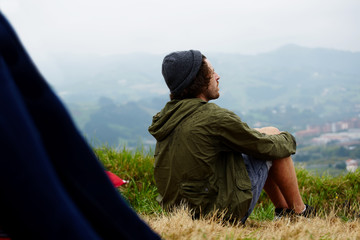 Fototapeta premium Young hike hipster man sitting at his campsite on the top of hill while enjoying the nature in rainy weather, young man relaxing on the grass enjoying outdoors view and relaxing after hike way