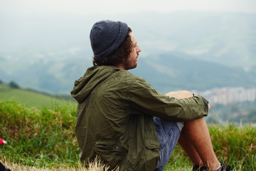 Young hike hipster man sitting at his campsite on the top of hill while enjoying the nature in rainy weather, young man relaxing on the grass enjoying outdoors view and resting after hike way