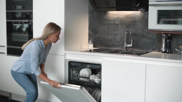 Young pretty woman unloading dishwasher in modern stylish kitchen