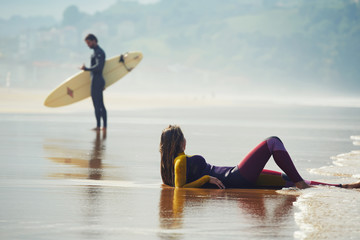 Young surfer standing on a beach holding a surfboard with a girl lying on sand on the foreground