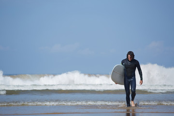 Young surfer sport man carrying his surfing board and going out the sea while on vacation during a sunny day, with an intense blue sky and big waves break on background