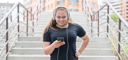 A fat young woman goes down the stairs and listens to music on a smartphone. Obese girl with...