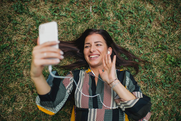 Fototapeta premium Young Caucasian woman lying down in the grass and taking selfie