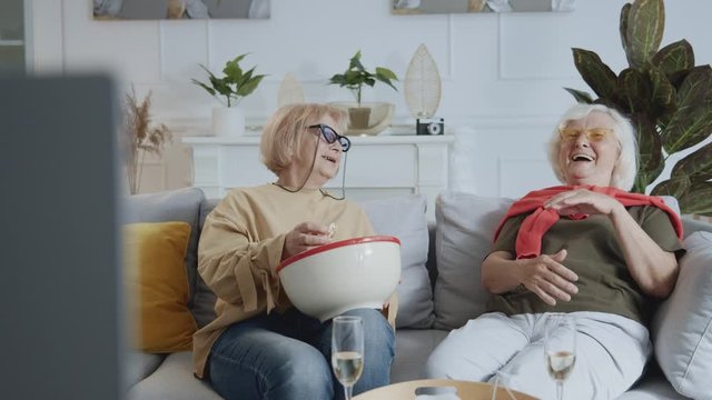 Joyful fashion grannies in luxury apartment watching TV movies eating pop cron laughing having good time. Entertainment. Best friends.