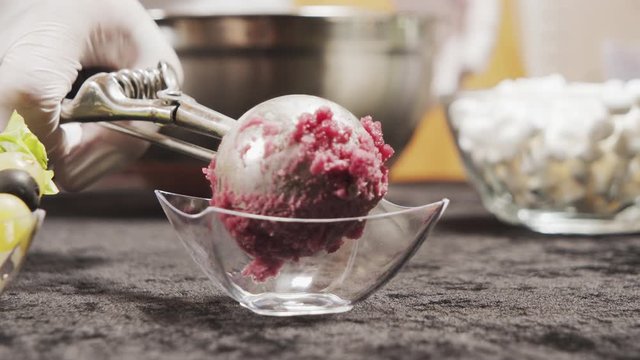 Close-up shot of delicious red wine ice cream ball with a piece of chocolate in a transparent bowl. Chef's hands in gloves serving a portion of ice cream.
